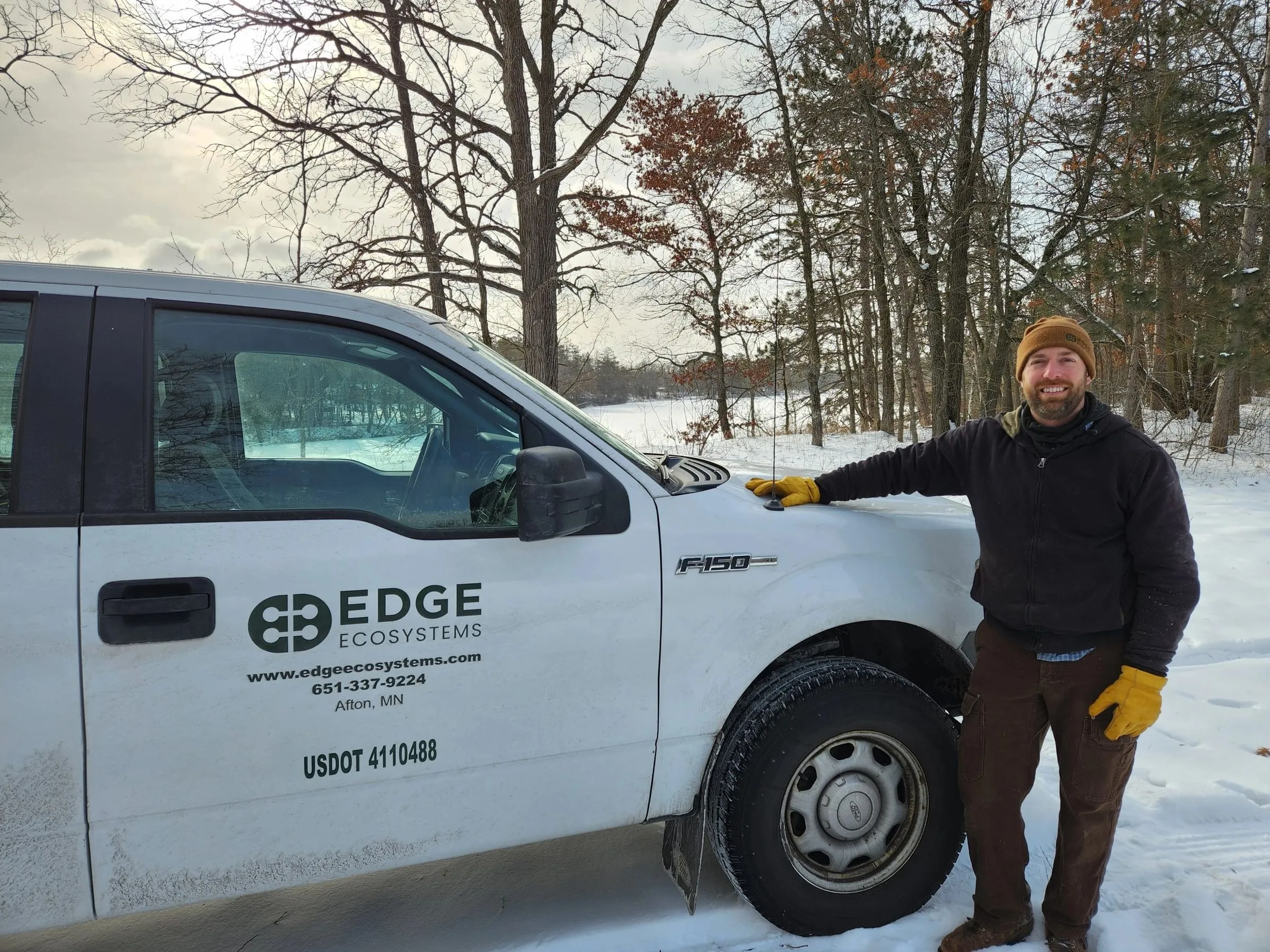 A member of the Edge Ecosystems crew in a knit cap and work gloves standing beside the company pickup on a snowy winter job site in an oak woodland