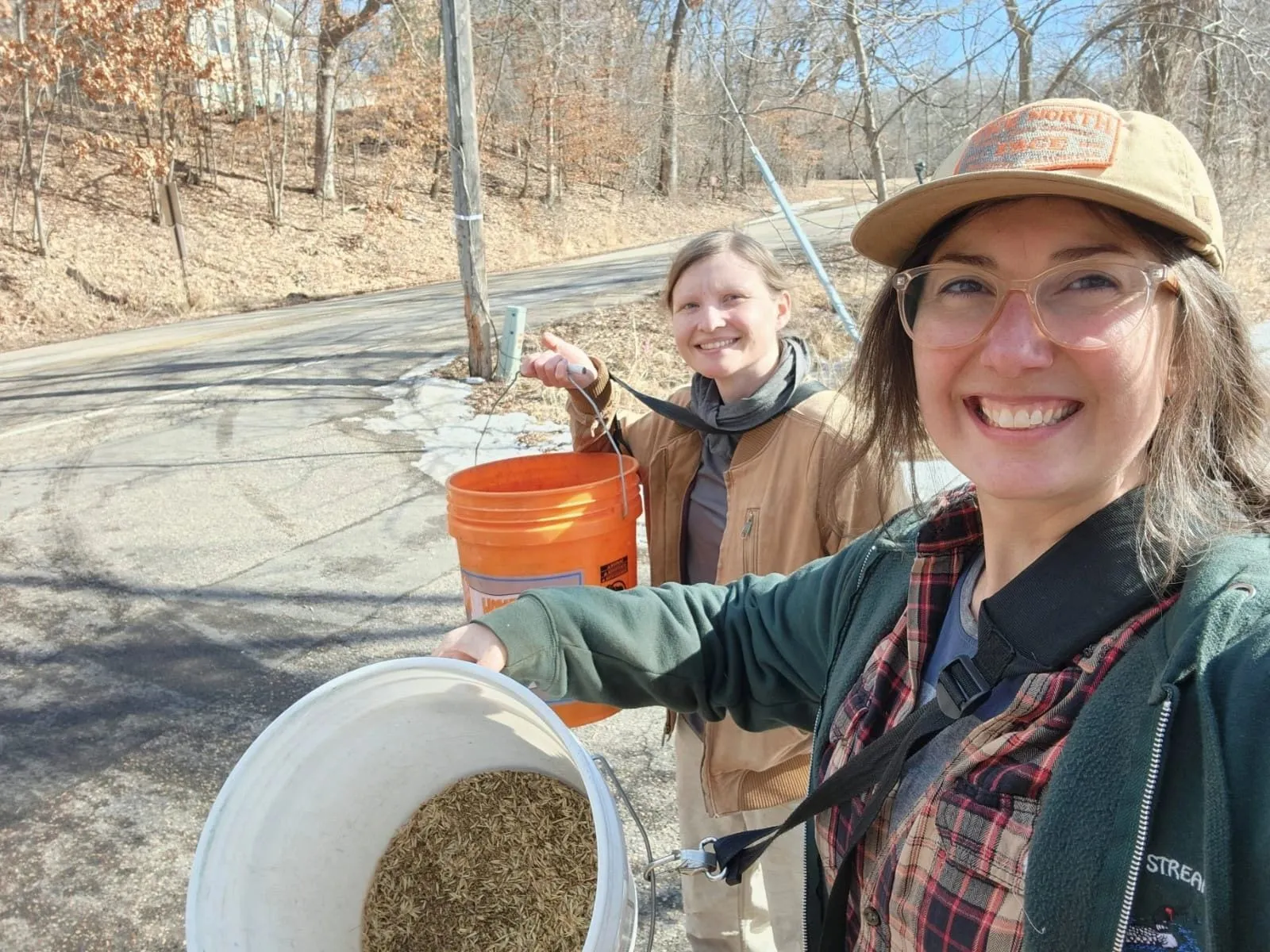 Two people on a roadside in late winter holding buckets of native seed, bare oaks and patchy snow behind them
