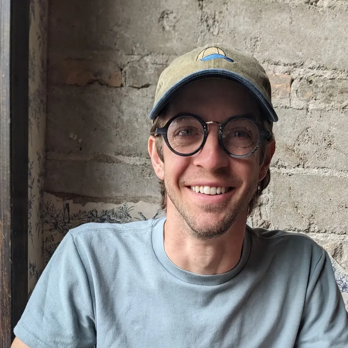 Alex Odden smiling in a canvas ball cap and round tortoiseshell glasses, standing against a weathered brick and plaster wall.